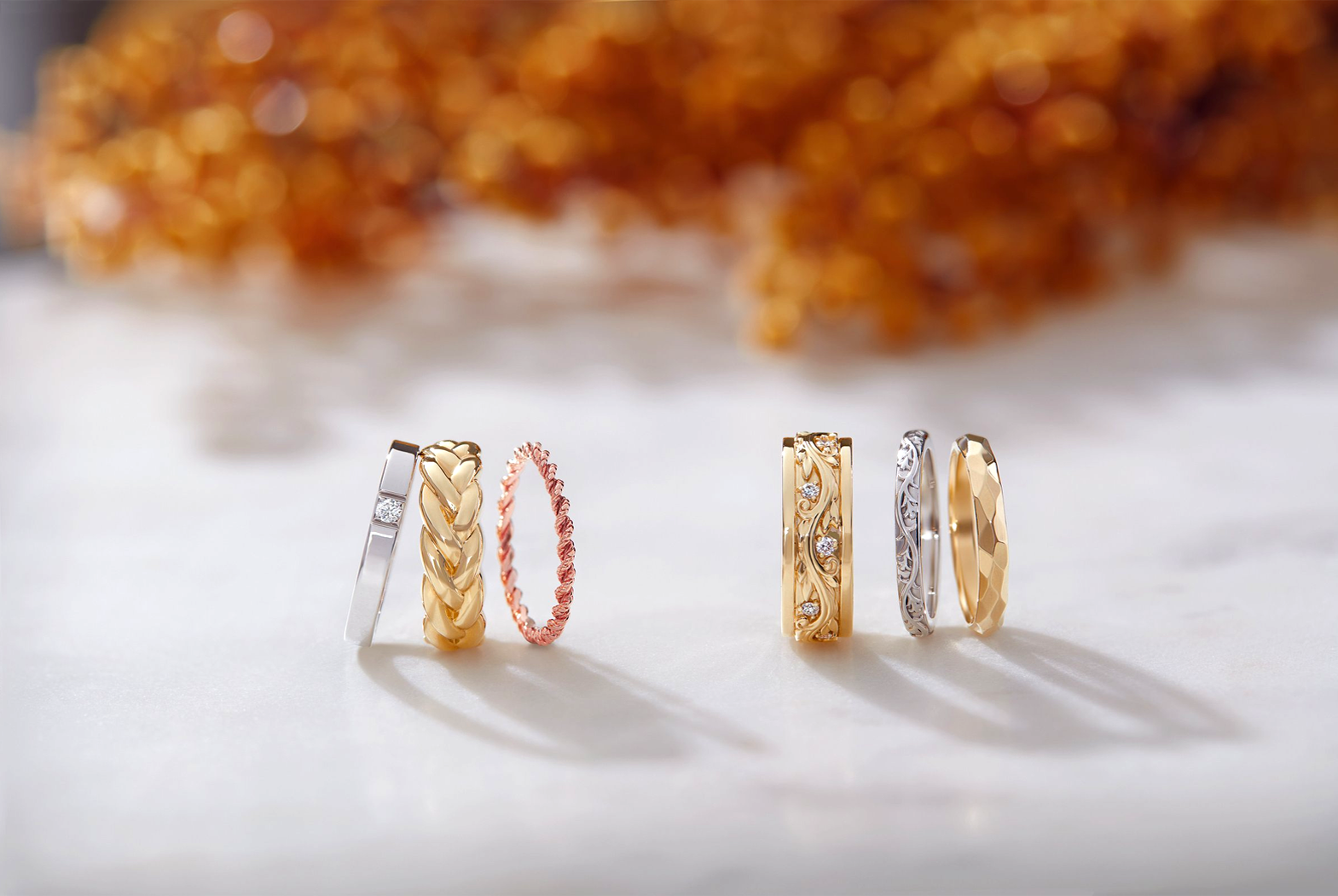 Set of gold and rose gold rings on a light surface with blurred orange flowers in the background